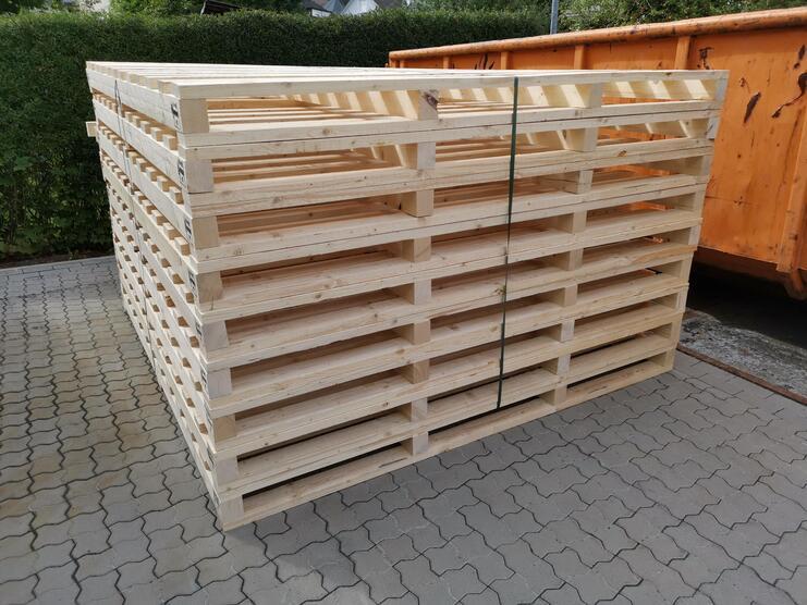 Stapel aus massiven Holzpaletten auf einem gepflasterten Boden, mit einem grünen Hintergrund und einem Abfallcontainer.